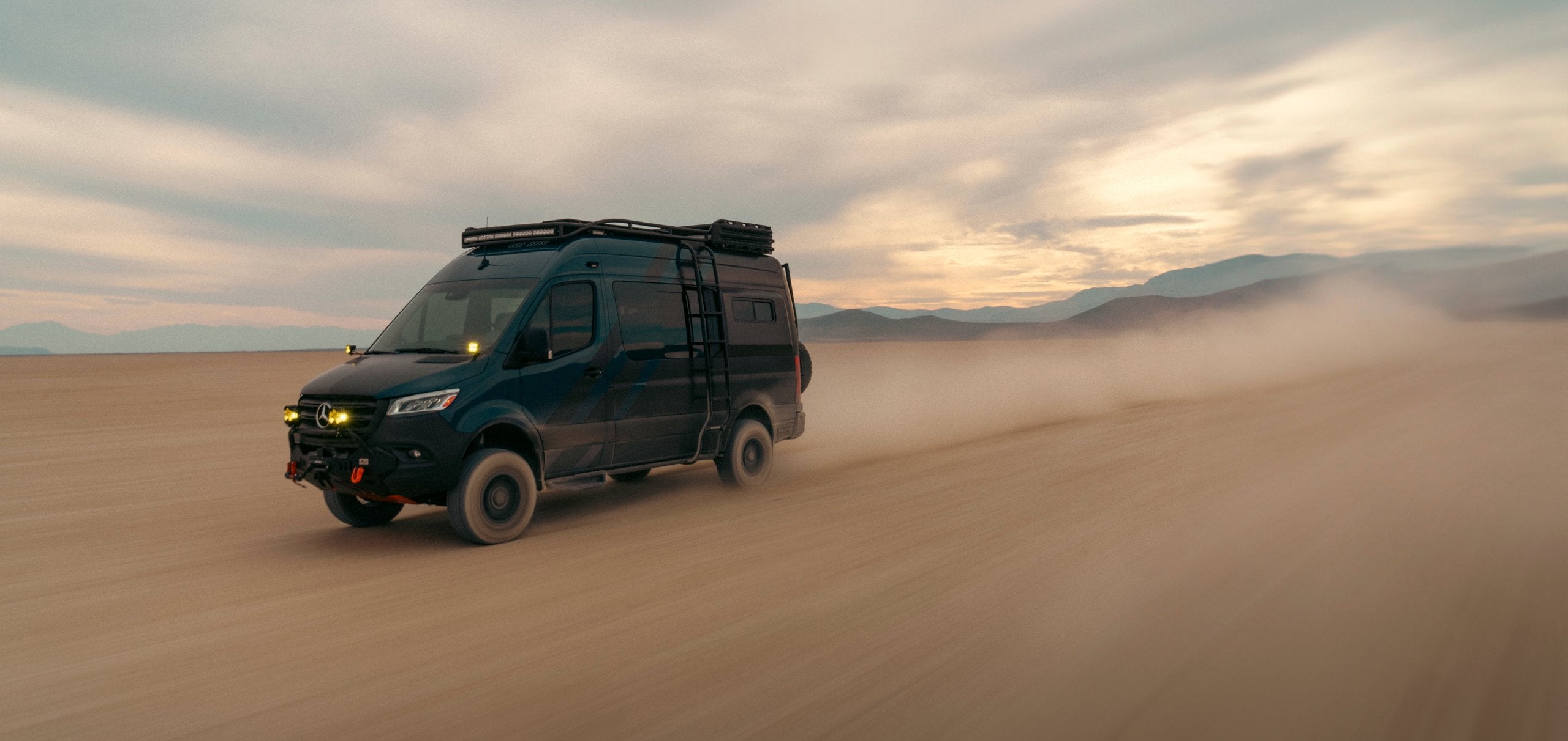 Mercedes Sprinter 4x4 with a Paragon Offroad roof rack and side ladder crossing a dry lake bed at dusk